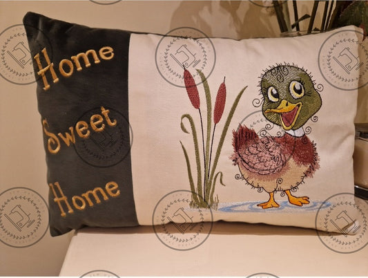 "An embroidered design of a cheerful mallard duck with a vibrant green head, brown and beige body, and yellow beak. The duck is standing in a small blue puddle next to tall cattail reeds, with a whimsical and curly detailing style."