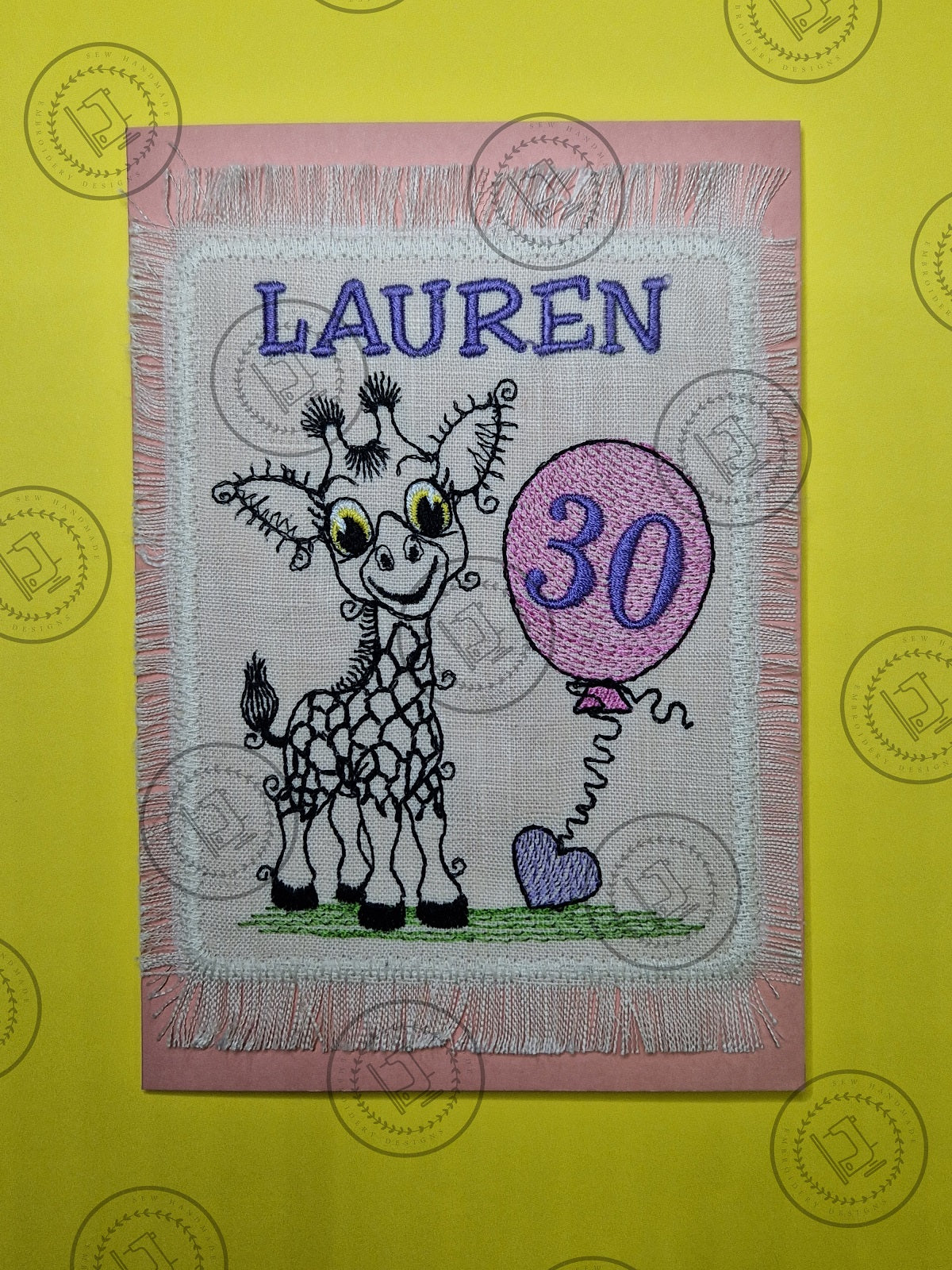 "An embroidered design of a whimsical giraffe with a cheerful expression, featuring exaggerated curly eyelashes and black-patterned spots. The giraffe is standing on a patch of green grass and holding a pink balloon with a curly string tied to a small purple heart."