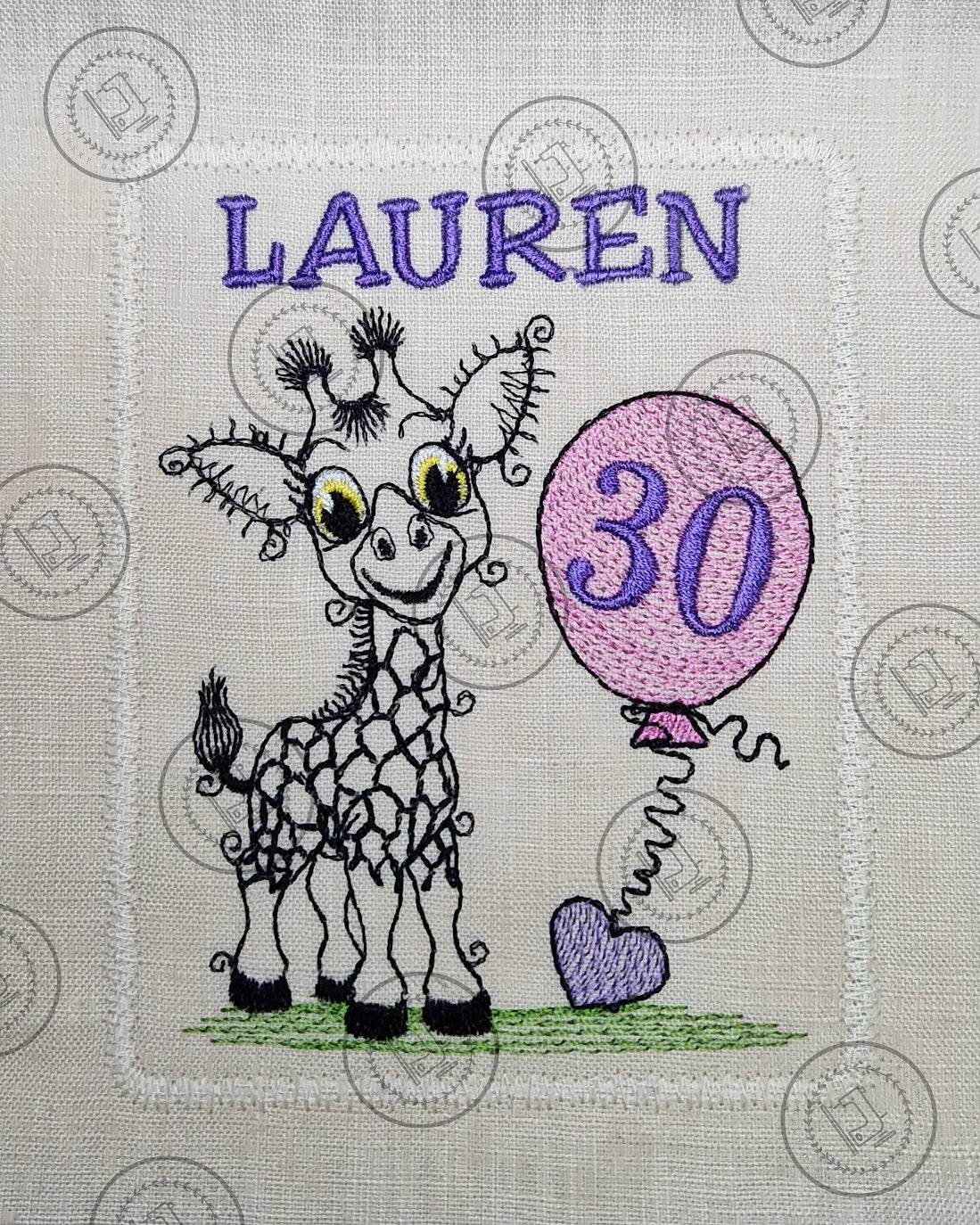 "An embroidered design of a whimsical giraffe with a cheerful expression, featuring exaggerated curly eyelashes and black-patterned spots. The giraffe is standing on a patch of green grass and holding a pink balloon with a curly string tied to a small purple heart."