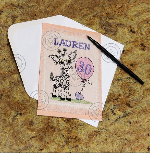 "An embroidered design of a whimsical giraffe with a cheerful expression, featuring exaggerated curly eyelashes and black-patterned spots. The giraffe is standing on a patch of green grass and holding a pink balloon with a curly string tied to a small purple heart."