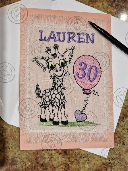 "An embroidered design of a whimsical giraffe with a cheerful expression, featuring exaggerated curly eyelashes and black-patterned spots. The giraffe is standing on a patch of green grass and holding a pink balloon with a curly string tied to a small purple heart."